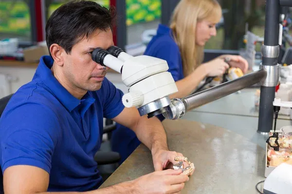 A dental technician verifying a restoration under a stereo microscope at the bench.